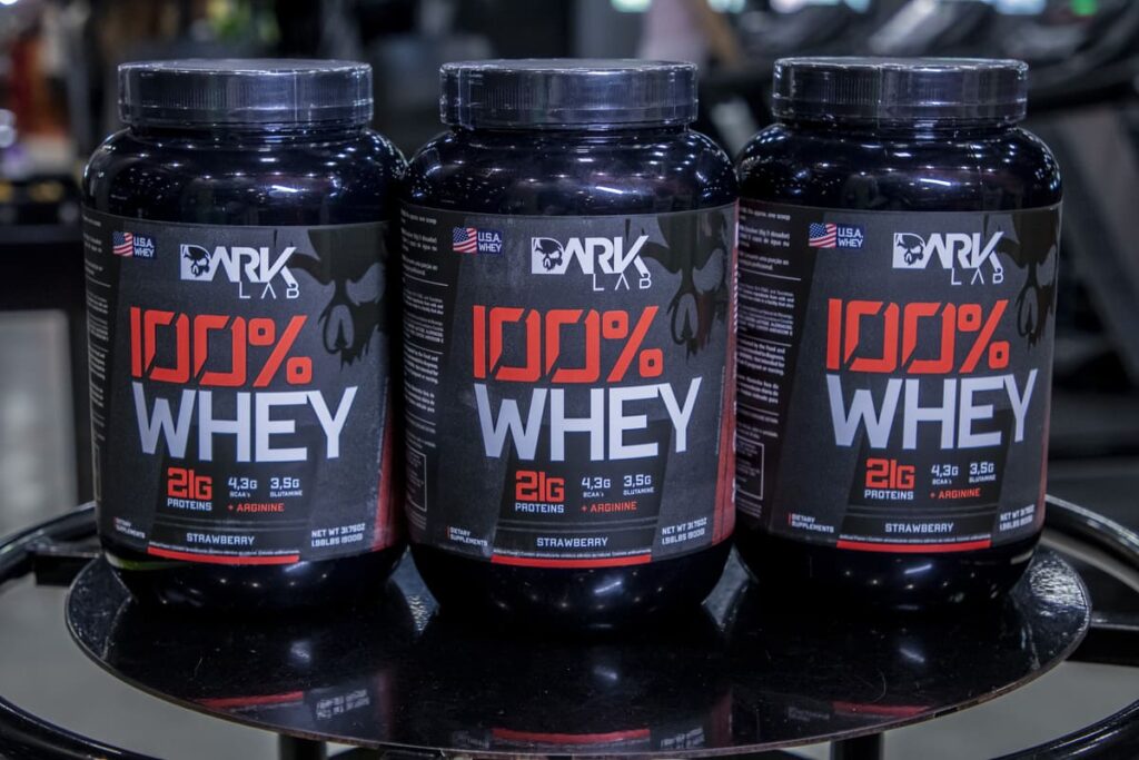 Três potes idênticos do suplemento alimentar 100% WHEY DARK LAB . O produto contém 21g de proteínas, 4,3g de BCAA e 3,5g de Arginina por porção, sendo um dos tipos de Whey Protein concentrado disponíveis no mercado.