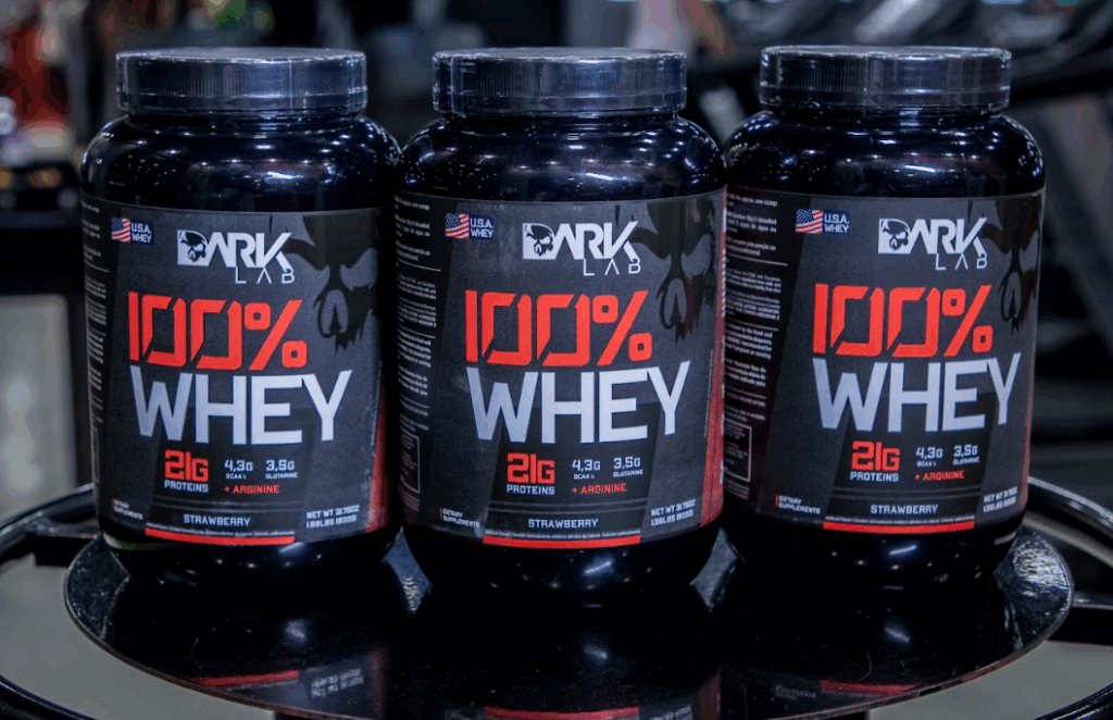 Tipos de suplementos: três potes de whey protein sabor morango da marca DARK Lab, exibidos em uma mesa redonda com tampo de vidro preta.