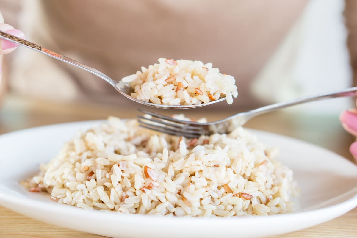 Close-up de arroz integral, um dos alimentos ricos em carboidratos, sendo servido com colher e garfo em um prato branco.