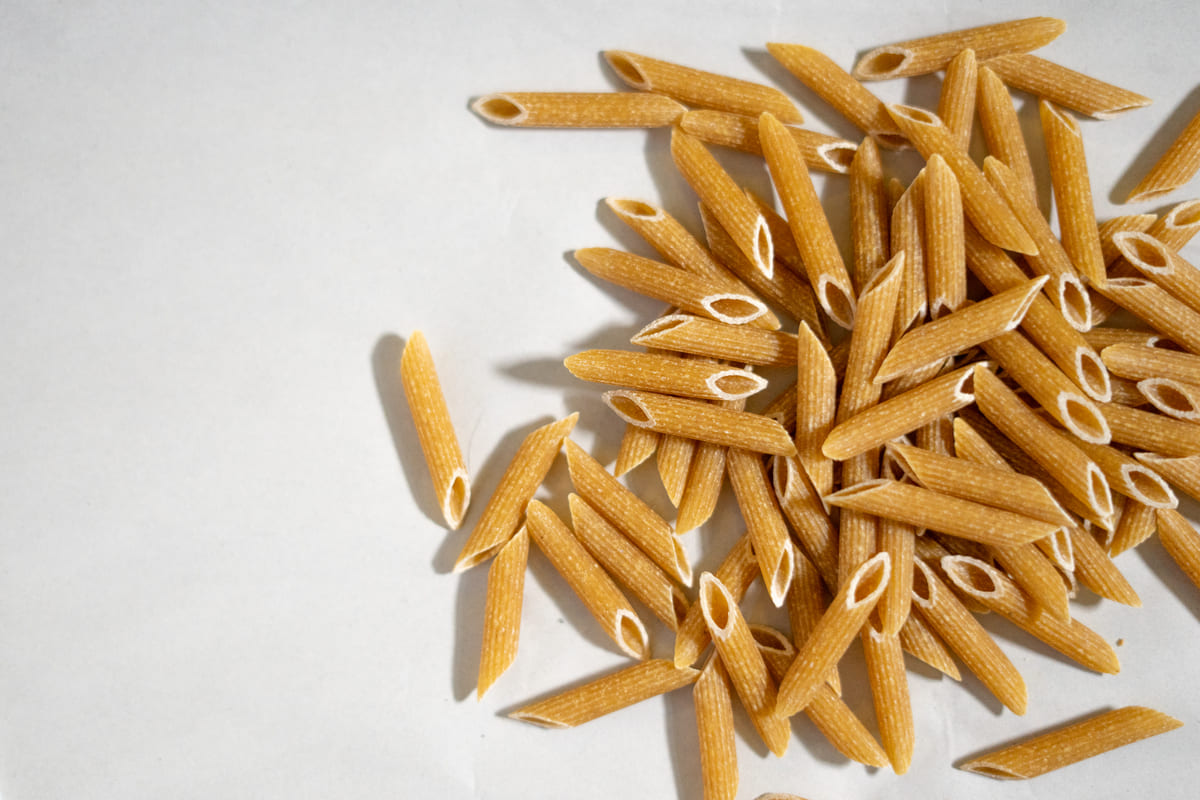 Macarrão penne integral cru, um dos alimentos ricos em carboidratos, espalhado no canto direito sobre uma superfície clara.