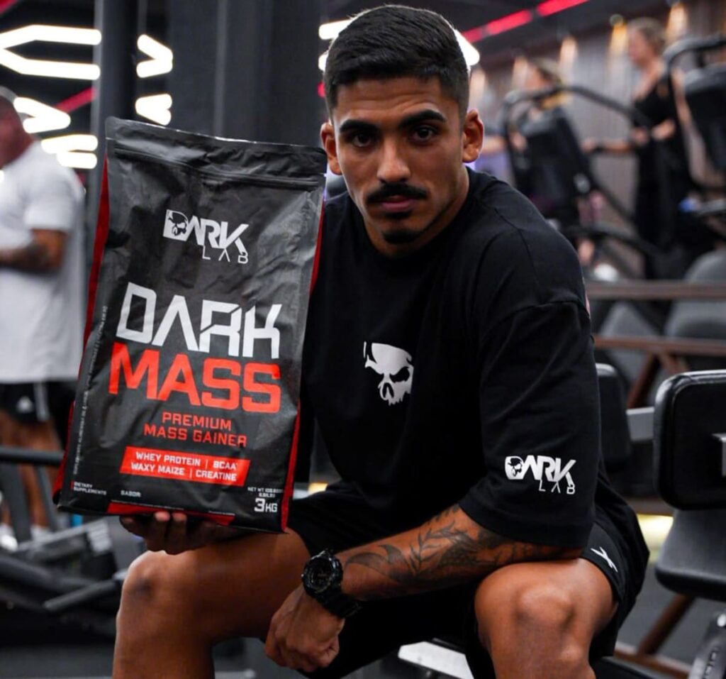 Homem em academia vestindo camiseta DARK LAB, segurando um pacote de Hipercalórico Dark Mass. O suplemento ajuda a ganhar massa muscular e a aumentar o gasto calórico diário, favorecendo a Taxa Metabólica Basal.