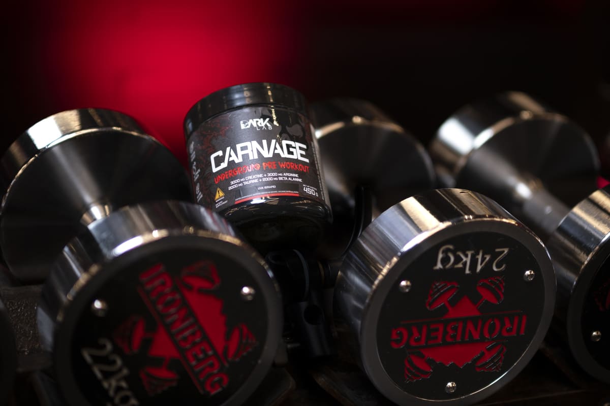 Pote do Pré-Treino Carnage Dark Lab posicionado entre halteres. O suplemento fornece a energia e o foco para o treino de powerlifting de alta intensidade.