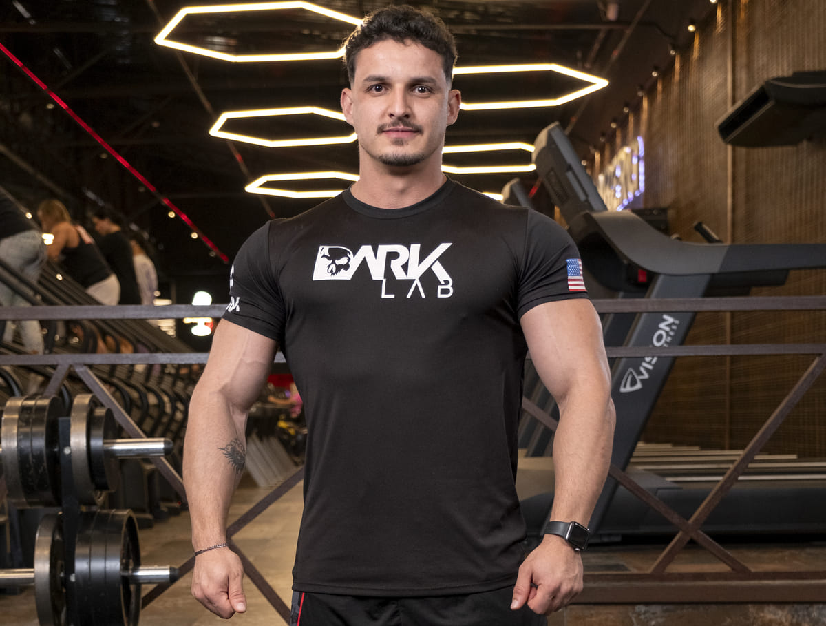 Atleta da DARK LAB em pé na academia, demonstrando foco e preparo físico. Manter o condicionamento e respeitar os intervalos de treino é essencial para evitar lesão muscular.