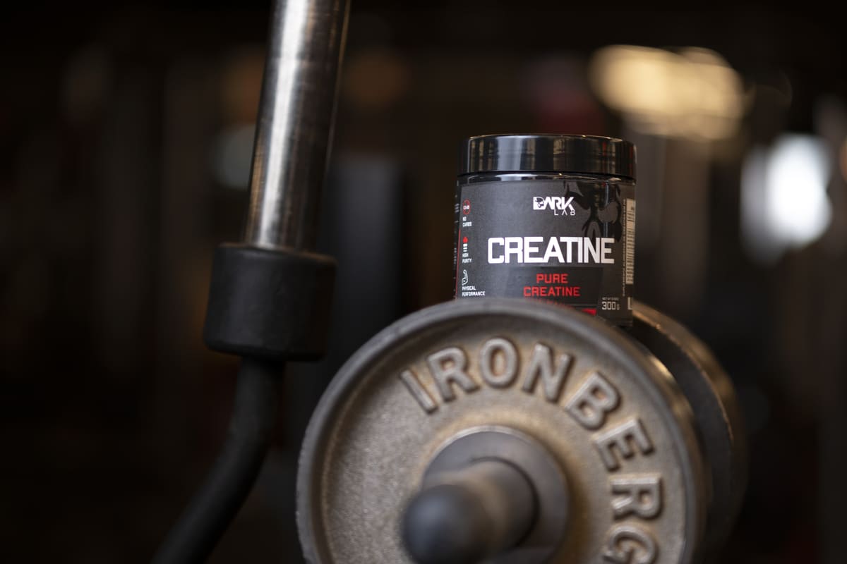 Pote de Creatina Pura DARK LAB sobre anilha de peso em ambiente de academia. A Creatina é fundamental para o ganho de força, a recuperação muscular e a prevenção de lesão muscular.